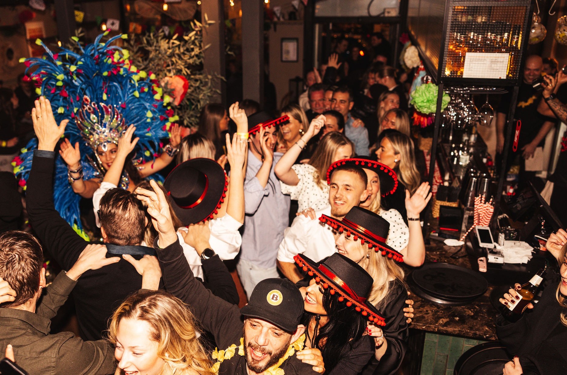 Fullsatt fiesta i baren med gäster i röda cordobés-hattar och sambafjädrar